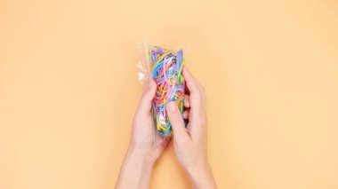 Ellerin bej arkaplanda renkli lastik bantlarla dolu temiz bir plastik poşet tutuş görüntüsü. El işi, DIY projeleri ve organizasyon için mükemmel. Eğlenceli ve enerjik..