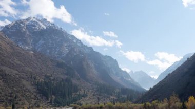 Açık mavi gökyüzünün altında karla kaplı dağlar. Engebeli arazi kayalık yamaçlara ve dağınık bulutlara sahip, sakin bir doğal manzara yaratıyor..
