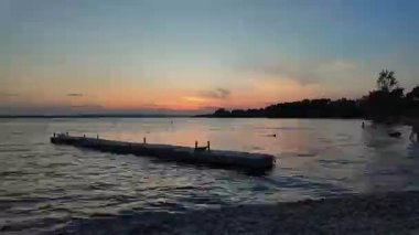 Sakin bir gölün üzerinde sakin bir günbatımı. Tahta bir rıhtım suya doğru uzanıyor. Ağaçların siluetleri kıyı boyunca uzanır. Gökyüzü sıcak turuncu ve mor renkler sergiliyor..