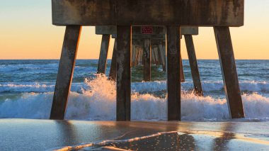 İskele yapısı, gün batımında düşen okyanus dalgalarının üzerinde yükselir. Islak kumu yansıtır ve renkli gökyüzü ile sakin bir sahne yaratır.