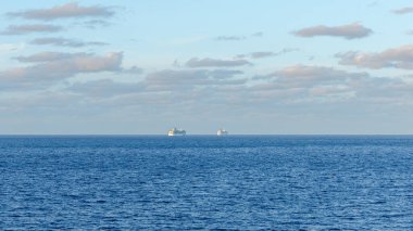 İki gemi, berrak bir gökyüzü ve kabarık bulutlarla çevrili, okyanusun sakin mavi sularında geziniyor. Huzurlu bir deniz manzarasını çağrıştırıyor.