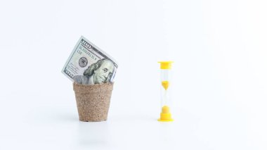 100 dolarlık banknot, zaman kavramının para olduğunu sembolize ederek kum saatinin yanında küçük bir tencerede duruyor. Bu görüntü yatırımı, finansal büyümeyi ve zamanlamanın önemini temsil ediyor..
