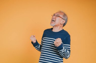 Zafer duygusunu kutlamak için yükselen yaşlı yaşlı adam kollarının heyecanlı ifadesi, gözlüklü mutlu Asyalı yaşlı adam evet sarı arka planda kazanan duygu gibi.