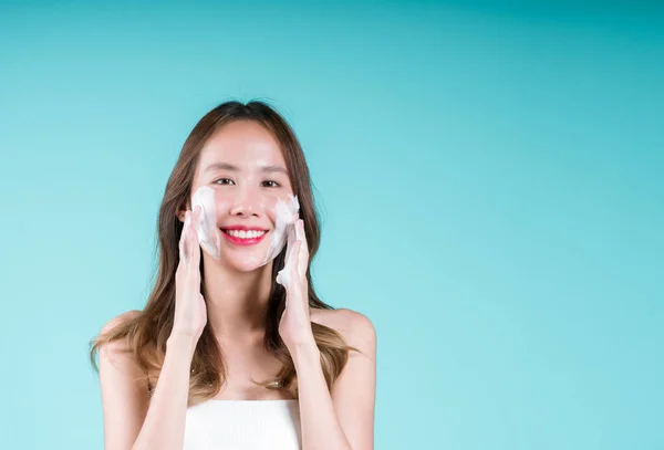 快乐迷人的年轻亚洲女人在蓝色背景的脸上用泡沫清洁剂洗净和擦拭 — 图库照片