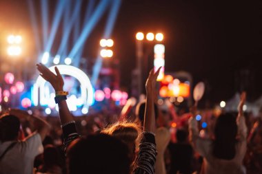 Kalabalık sahne ışıklarını yakıyor ve insanlar seyirci siluetini hayran bırakıyor. Müzik festivalinin arka planında sahne ışığı gece hayatı etkinliğiyle el kaldırıyor..