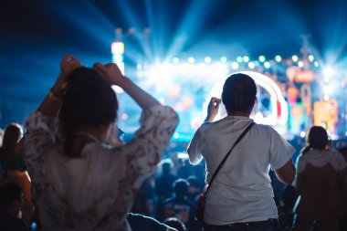 Kalabalık sahne ışıklarını yakıyor ve insanlar seyirci siluetini hayran bırakıyor. Müzik festivalinin arka planında sahne ışığı gece hayatı etkinliğiyle el kaldırıyor..