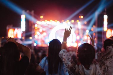 Kalabalık sahne ışıklarını yakıyor ve insanlar seyirci siluetini hayran bırakıyor. Müzik festivalinin arka planında sahne ışığı gece hayatı etkinliğiyle el kaldırıyor..