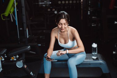 Kadın saati kontrol ediyor ve spor salonunda egzersiz yapıyor. Spor yaptıktan sonra halterler ve su şişesiyle vücut geliştirme egzersizleri yapıyor..