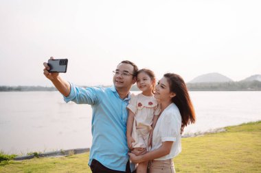 Hafta sonu parkta mutlu bir aile. Nehir kenarındaki çayırda birlikte eğlenen aile kızı tutuyor. Sağlık sigortası konsepti.