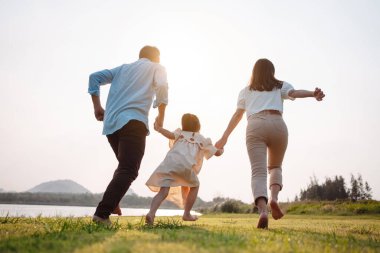 Parkta gün batımında mutlu bir aile. Aile hafta sonu çayırda birlikte koşarken nehir aileleri çocuğun elini tutuyor. Sağlık sigortası kavramı..