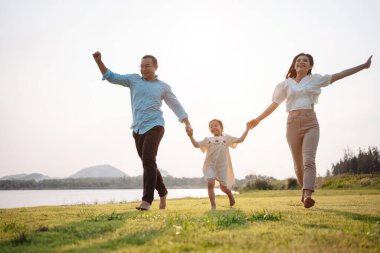 Parkta gün batımında mutlu bir aile. Aile hafta sonu çayırda birlikte koşarken nehir aileleri çocuğun elini tutuyor. Sağlık sigortası kavramı..