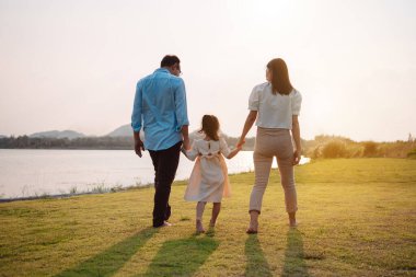Parkta gün batımında mutlu bir aile. Aile hafta sonu çayırda birlikte koşarken nehir aileleri çocuğun elini tutuyor. Sağlık sigortası kavramı..