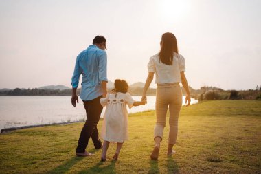 Parkta gün batımında mutlu bir aile. Aile hafta sonu çayırda birlikte koşarken nehir aileleri çocuğun elini tutuyor. Sağlık sigortası kavramı..
