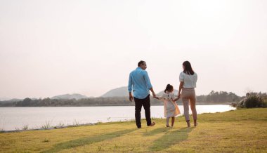 Parkta gün batımında mutlu bir aile. Aile hafta sonu çayırda birlikte koşarken nehir aileleri çocuğun elini tutuyor. Sağlık sigortası kavramı..