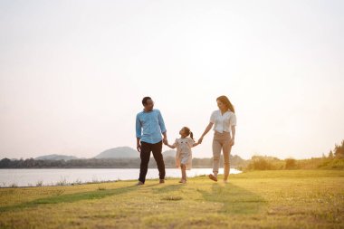 Parkta gün batımında mutlu bir aile. Aile hafta sonu çayırda birlikte koşarken nehir aileleri çocuğun elini tutuyor. Sağlık sigortası kavramı..