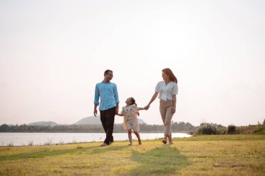 Parkta gün batımında mutlu bir aile. Aile hafta sonu çayırda birlikte koşarken nehir aileleri çocuğun elini tutuyor. Sağlık sigortası kavramı..