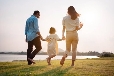 Parkta gün batımında mutlu bir aile. Aile hafta sonu çayırda birlikte koşarken nehir aileleri çocuğun elini tutuyor. Sağlık sigortası kavramı..