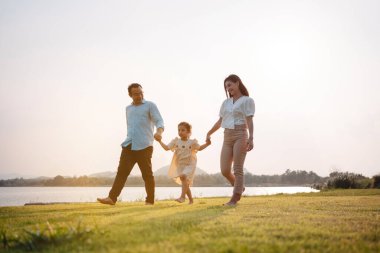 Parkta gün batımında mutlu bir aile. Aile hafta sonu çayırda birlikte koşarken nehir aileleri çocuğun elini tutuyor. Sağlık sigortası kavramı..