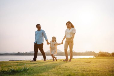 Parkta gün batımında mutlu bir aile. Aile hafta sonu çayırda birlikte koşarken nehir aileleri çocuğun elini tutuyor. Sağlık sigortası kavramı..