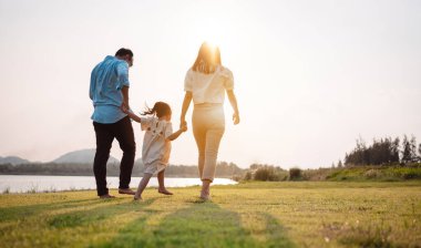 Parkta gün batımında mutlu bir aile. Aile hafta sonu çayırda birlikte koşarken nehir aileleri çocuğun elini tutuyor. Sağlık sigortası kavramı..