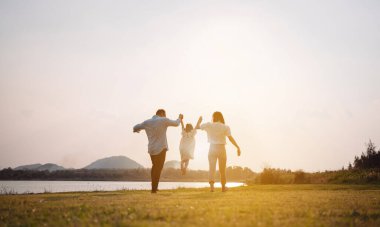 Parkta gün batımında mutlu bir aile. Aile hafta sonu çayırda birlikte koşarken nehir aileleri çocuğun elini tutuyor. Sağlık sigortası kavramı..