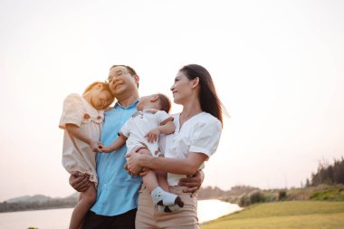 Parkta gün batımında mutlu bir aile. Aile hafta sonu çayırda birlikte oynuyorlardı.Nehir kenarındaki ebeveynler çocuk ve kızın ellerini tutuyorlar. Sağlık hayat sigortası konsepti.