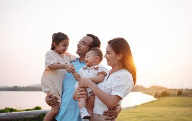 Parkta gün batımında mutlu bir aile. Aile hafta sonu çayırda birlikte oynuyorlardı.Nehir kenarındaki ebeveynler çocuk ve kızın ellerini tutuyorlar. Sağlık hayat sigortası konsepti.