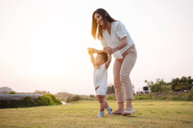 Bebek gün batımında çayırda ilk adımlarını atıyor. Anne, çocuğun ileri doğru yürümesini, bebek büyümesini ve gelişmesini destekliyor.