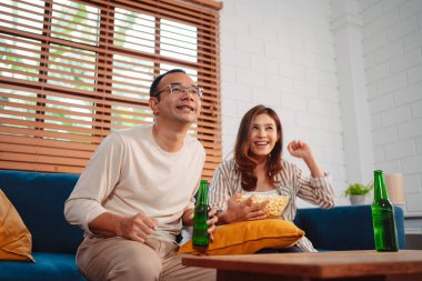 Asyalı çift, evdeki oturma odasındaki kanepede maç izlerken sevinçten havalara uçuyor. Rahatlatıcı mutluluğun tadını çıkar.