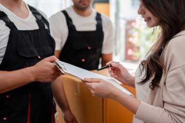 Kadın, kutuları ve mobilyaları bir kamyonetten yeni bir eve taşırken kontrol et. Üniformalı iki erkek nakliyeci, kutuları kaldırıyor. Ev Taşıma ve Teslim Konsepti.