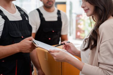 Kadın, kutuları ve mobilyaları bir kamyonetten yeni bir eve taşırken kontrol et. Üniformalı iki erkek nakliyeci, kutuları kaldırıyor. Ev Taşıma ve Teslim Konsepti.