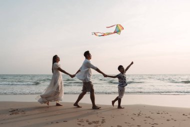 Mutlu Asyalı aile deniz sahilinin tadını çıkarıyor. Baba, anne ve oğul yaz tatilinde sahilde uçurtma oynarken eğleniyorlar. Okyanus kıyısında tatil zamanı yaşam tarzı konsepti..