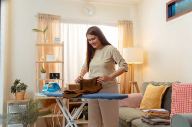 Ev işi yapan kadın ev işlerini ütülüyor ev işi ev eşyaları asyalı kadın teçhizatı, ütü yaparken ve katlarken gülümseyen kadın temiz bir oturma odasında, neşeli bir ev rutinini kucaklıyor