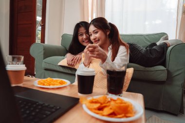 Asyalı Kadın Atıştırmalığı Yaşam Tarzı Oturma Odası Keyif Filmi, Televizyon izlerken İki Genç Yetişkin Kahkaha ve Atıştırma, Evde Arkadaşlık ve Gevşemeyi Kucaklaşma