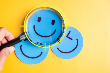 Duygusal değerlendirme ve olumlu zihniyet konsepti. Büyüteç, birden fazla mutlu ifadeyle gülen yüze odaklanıyor. Ruh sağlığı, geri bildirim, ruh hali kontrolü ve psikolojik bakım için kullanışlı.