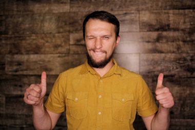 Sakallı, sarı tişörtlü, genç, yakışıklı ve yakışıklı bir adam. Ahşap arka planda olumlu bir jest yapmayı onaylıyor. Gülümsüyor ve başarı için seviniyor. Kazanan jest