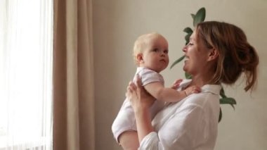 Sevgi dolu genç bir anne kucağında sevimli bir kız bebek kucağında küçük bir çocuğa sarılıyor. Mutlu şefkatli bir anne, evde pencerenin önünde duran küçük çocuğa sarılıyor. Mutlu annelik kavramı.