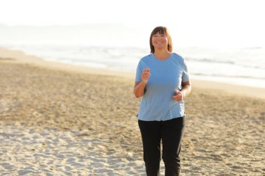 Gün doğumunda kumsalda koşan gülümseyen orta yaşlı kadın. 40 'lı ya da 50' li yaşlarda, spor kıyafetleri içinde, spor yapan, spor yapan, spor yapan, güzel deniz manzarasında sağlıklı yaşam tarzını ve sporu seven olgun bir kadın.