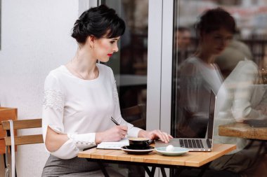 Genç iş kadını dizüstü bilgisayarda çalışıyor ve şehir merkezindeki terastaki kafede not defterine notlar yazıyor. Beyaz bluzlu bir kıyafet. Kahve molası, iş konsepti. Açık havada çalışmak. Uzak görev
