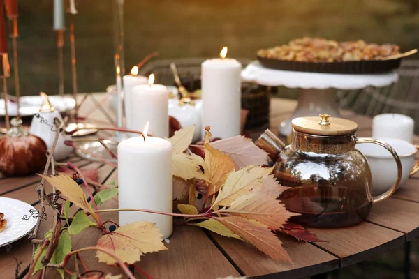 Cozy autumn picnic in the park. Close up of glass kettle with hot black tea, classic cups, homemade apple pie, maple leaves and burning candles on wooden table outdoors