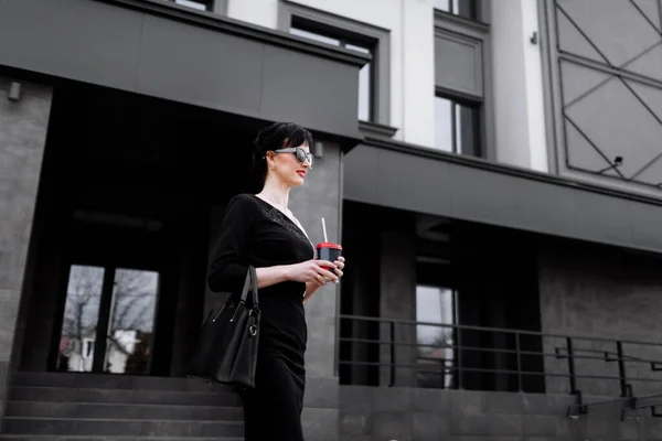 Smiling business woman in sunglasses walks down the business central city street with offices and holding cup of coffee and has break. Pretty woman in has a business dress code and wearing in dress.