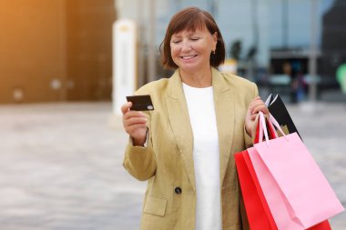 Alışveriş merkezinin arka planında duran kredi kartı, banka kartı ve alışveriş çantalarıyla gülümseyen şık, orta yaşlı bir kadın. Sonbahar tatilleri, indirimler, Kara Cuma. Tüketim, satış, zengin hayat kavramı.