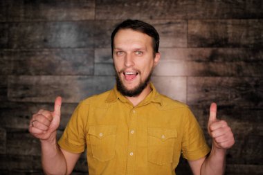 Sakallı, sarı tişörtlü, genç, yakışıklı ve yakışıklı bir adam. Ahşap arka planda olumlu bir jest yapmayı onaylıyor. Gülümsüyor ve başarı için seviniyor. Kazanan jest