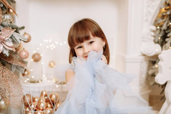 お祝いのプリンセスブルーのドレスで愛らしい笑顔の女の子は居心地の良い部屋の照明ガーランド装飾のカメラで探しています 幸せな家族の休日 家で居心地の良い暖かい冬の夜 — ストック写真