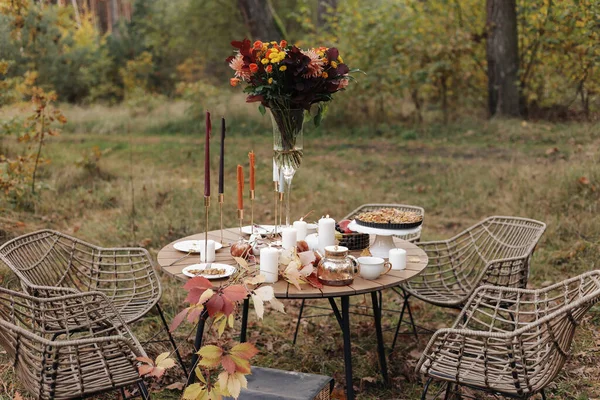 Cozy autumn picnic in the park. Festive setting table decorated wildflowers in vase, candles, food, hot drinks, maples leaves and wicker chairs outdoors. Straw baskets and pumpkins on the grass.