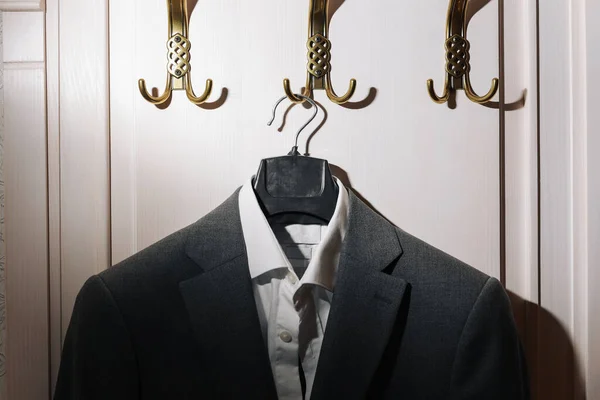 男のダークグレーのスーツジャケットと白いシャツは 白い壁の背景に金の金属フックにかかって — ストック写真