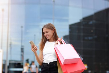 Kara Cuma. Alışveriş merkezinin arka planında, alışveriş sürecinde, tüketim, satış, zengin hayat kavramında, alışveriş poşetleriyle gülümseyen genç bir kadın akıllı telefon kullanıyor. Reklam için ticari teklif.