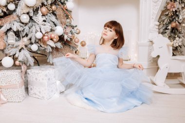 Şenlikli prenses mavisi elbisesi içinde sevimli gülümseyen küçük bir kız süslü çelenklerle dolu rahat bir odada kameraya bakıyor. Mutlu aile tatilleri, yeni yıl. Evde sıcak bir kış akşamı.