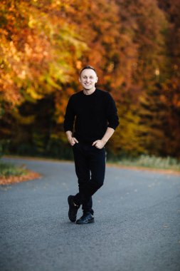 Genç çekici adam sonbahar ormanında yolda duruyor. Siyahlı kısa saçlı adam parktaki sarı yapraklı kameraya bakıyor. Kırsal kesimde sıcak bir gün.