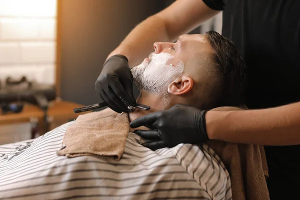 stock image Professional barber in black gloves is shaving client with a vintage straight stainless steel razor in barbershop. Shaving cream, foam. Hairdresser, beauty and fashion salon.